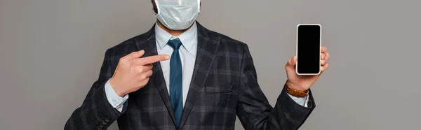 Обрізаний вид бізнесмена в медичній масці, вказуючи пальцем на смартфон з порожнім екраном ізольовано на сірому, панорамному посіві — стокове фото