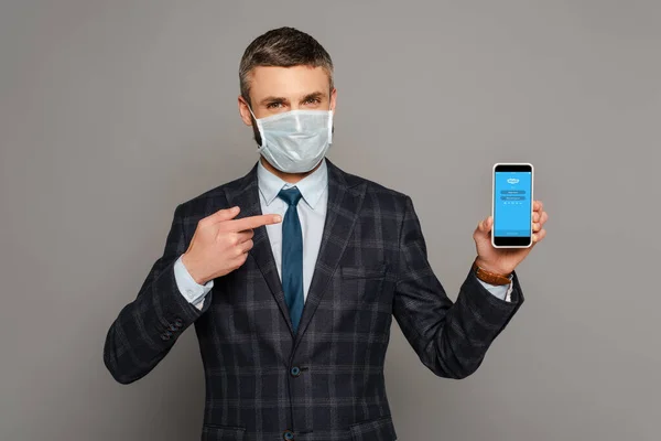 KYIV, UCRAINA - 30 MARZO 2020: bell'uomo d'affari in maschera medica che punta il dito contro smartphone con app skype su sfondo grigio — Foto stock