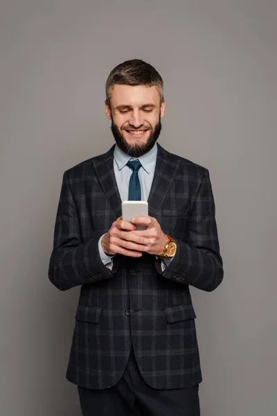 Homem de negócios barbudo bonito feliz em terno usando smartphone em fundo cinza — Fotografia de Stock