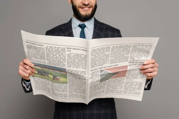 Vue recadrée du beau homme d'affaires barbu souriant en costume lisant le journal sur fond gris — Photo de stock