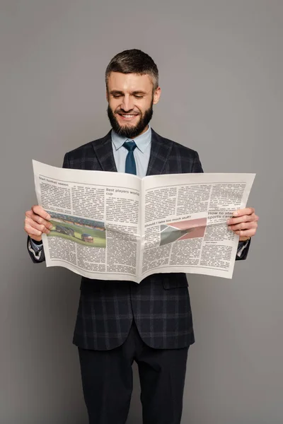 Smiling handsome bearded businessman in suit reading newspaper on grey background — Stock Photo