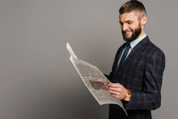 Smiling handsome bearded businessman in suit reading newspaper on grey background — Stock Photo