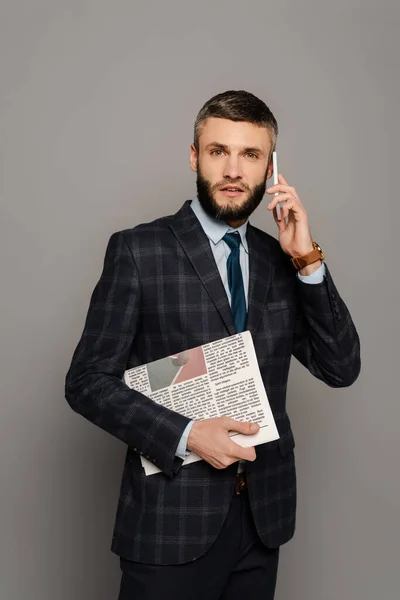 Handsome bearded businessman in suit with newspaper talking on smartphone on grey background — Stock Photo