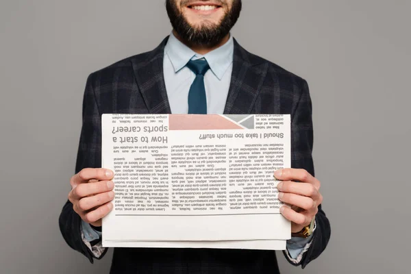Обрізаний вид усміхненого бородатого бізнесмена в костюмі з газетою на сірому фоні — стокове фото