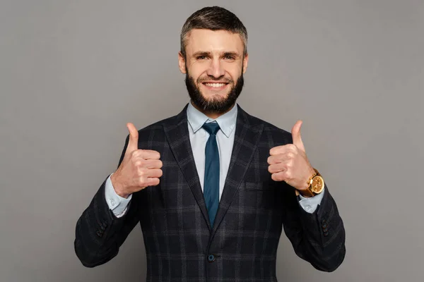 Feliz guapo barbudo hombre de negocios en traje mostrando pulgares hacia arriba sobre fondo gris - foto de stock