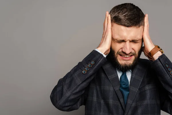 Guapo barbudo hombre de negocios en traje cubriendo las orejas con las manos sobre fondo gris - foto de stock