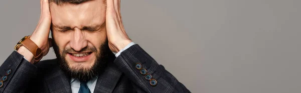 Guapo barbudo hombre de negocios en traje cubriendo las orejas con las manos aisladas en gris, cultivo panorámico - foto de stock