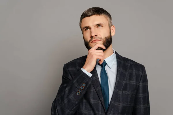 Nachdenklich schöner bärtiger Geschäftsmann im Anzug berührt Kinn auf grauem Hintergrund — Stockfoto