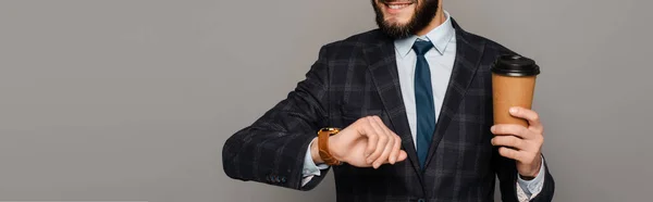 Smiling handsome bearded businessman in suit with coffee to go looking at wristwatch on grey background, panoramic orientation — Stock Photo