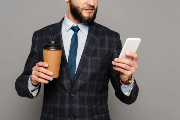 Vista cortada de empresário barbudo em terno com café para ir usando smartphone em fundo cinza — Fotografia de Stock