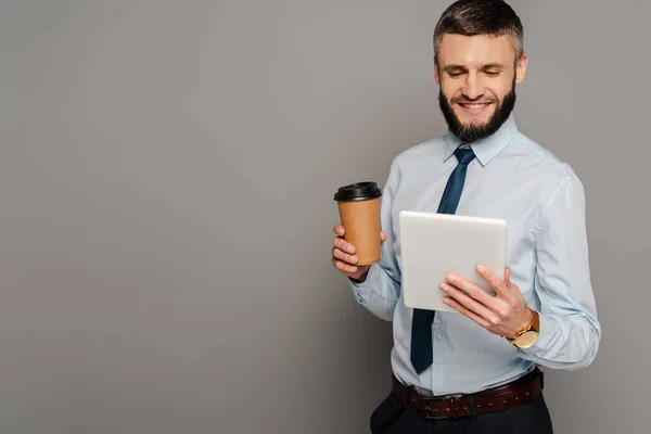 Homme d'affaires barbu beau sourire avec tablette numérique et café pour aller sur fond gris — Photo de stock