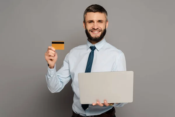 Sonriente hombre de negocios barbudo guapo con ordenador portátil y tarjeta de crédito sobre fondo gris - foto de stock