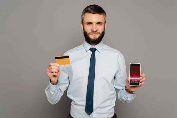 Handsome bearded businessman holding credit card and smartphone with trading courses app on grey background — Stock Photo
