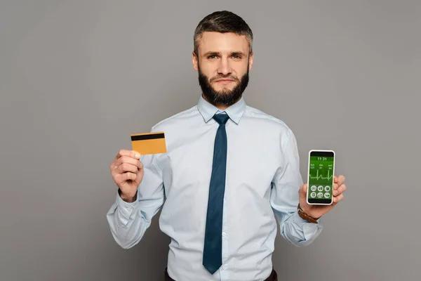 Guapo barbudo hombre de negocios con tarjeta de crédito y teléfono inteligente con aplicación de salud sobre fondo gris - foto de stock