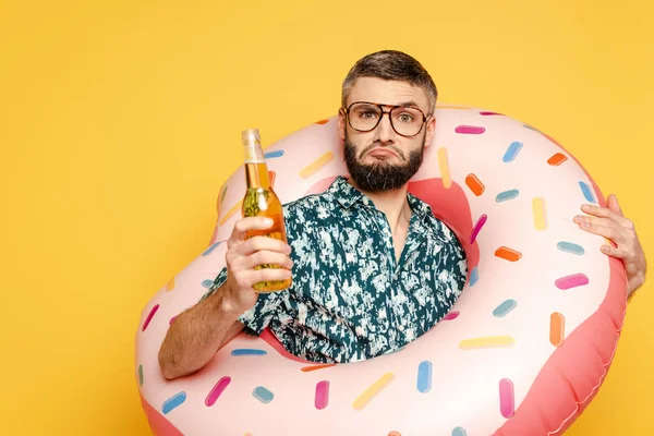 Сумний бородатий хлопець в окулярах і плавати кільце з пляшкою пива на жовтому — стокове фото
