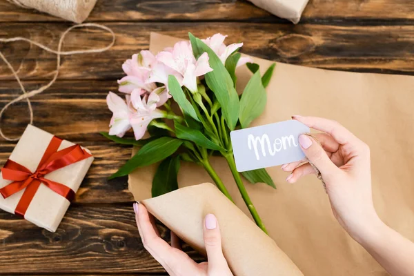 Top view of woman holding tag with mom lettering near pink flowers and gift box on wooden surface, mothers day concept — Stock Photo