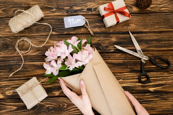 Cropped view of woman touching wrapped in paper pink flowers near gift boxes and tag with mom lettering on wooden surface, mothers day concept — Stock Photo