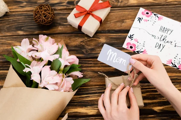 Vista ritagliata della donna toccando tag con scritta mamma sulla confezione regalo vicino a fiori rosa e biglietto di auguri con felice giorno delle madri sulla superficie di legno — Foto stock