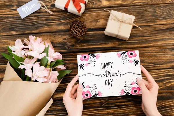 Vista recortada de la mujer que sostiene la tarjeta de felicitación con letras feliz día de las madres cerca de flores y regalos en la superficie de madera - foto de stock