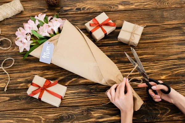 Vista recortada de la mujer cortando cuerda de yute en flores rosadas envueltas en papel cerca de cajas de regalo y tijeras, concepto de día de las madres - foto de stock