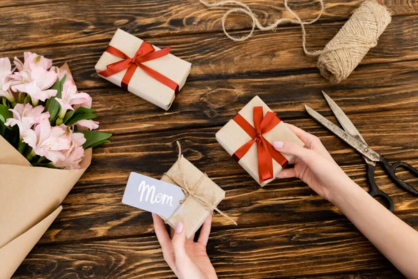 Cropped view of woman holding gift boxes near pink flowers wrapped in paper and jute twine rope on wooden surface, mothers day concept — Stock Photo