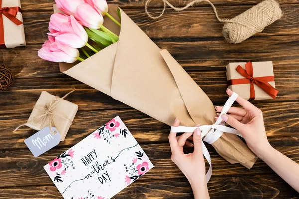 Vista cortada de mulher tocando fita em embrulhado em papel tulipas rosa perto de cartão de saudação com feliz dia das mães lettering e caixas de presente na superfície de madeira — Fotografia de Stock