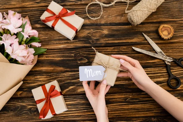 Cropped view of woman holding gift box with mom lettering on tag near pink flowers wrapped in paper on wooden surface, mothers day concept — Stock Photo