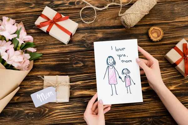 Vue recadrée de la femme tenant la carte de vœux avec je t'aime maman lettrage près de cadeaux et fleurs sur la surface en bois — Photo de stock