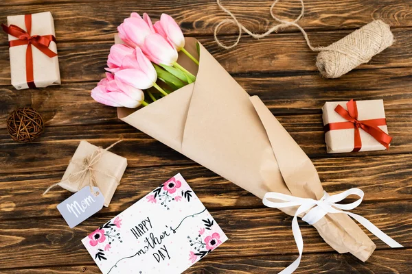 Vista superior de la tarjeta de felicitación con letras felices del día de las madres cerca de regalos y flores en la superficie de madera - foto de stock