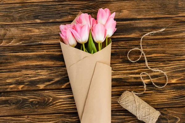 Top view of pink tulips wrapped in paper near jute twine rope on wooden surface, mothers day concept — Stock Photo