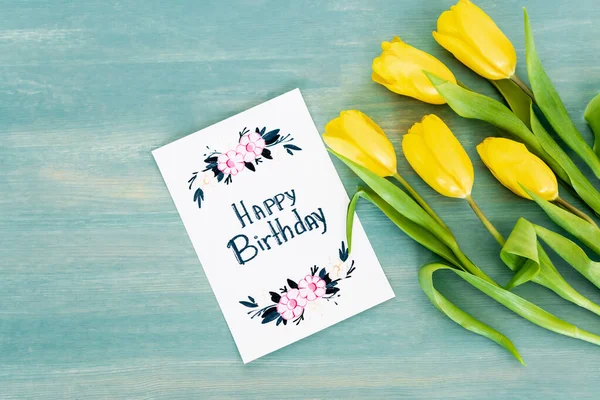 Top view of greeting card with happy birthday lettering near yellow tulips on blue textured surface — Stock Photo