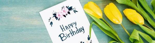 Panoramic shot of greeting card with happy birthday lettering near yellow tulips on blue textured surface — Stock Photo