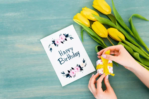 Cropped view of woman holding gift box near greeting card with happy birthday lettering and yellow tulips on blue wooden surface — Stock Photo