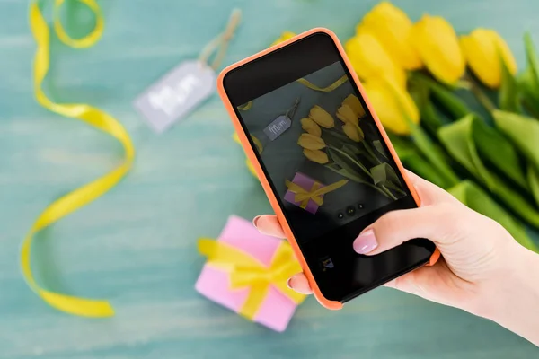 Selective focus of woman taking photo of yellow tulips, gift box and mom tag lettering  on textured surface, mothers day concept — Stock Photo