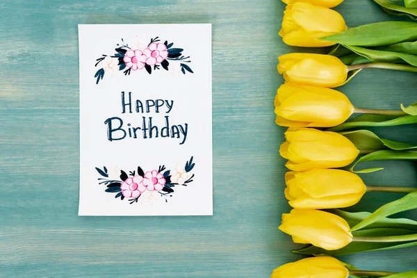 Top view of yellow tulips near greeting card with happy birthday lettering on blue textured surface — Stock Photo