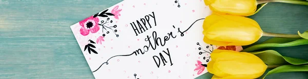 Panoramic shot of greeting card with happy mothers day lettering near yellow tulips on blue textured surface — Stock Photo