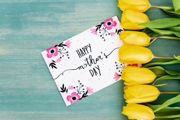 Top view of greeting card with happy mothers day lettering near yellow tulips on blue textured surface — Stock Photo