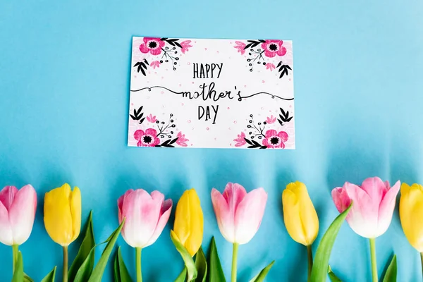 Top view of yellow and pink tulips near greeting card with happy mothers day lettering on blue — Stock Photo