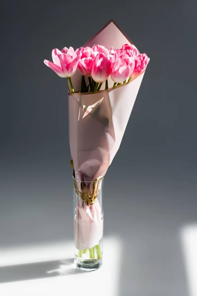 Lumière du soleil sur les tulipes roses et en fleurs dans un vase avec de l'eau sur gris, concept de fête des mères — Photo de stock