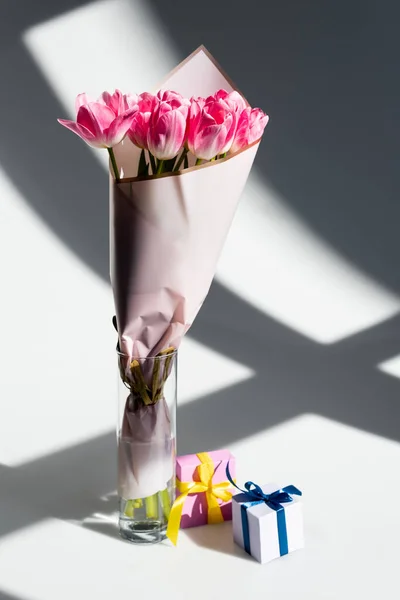 Luz del sol en tulipanes rosados en jarrón cerca de cajas de regalo en blanco, concepto de día de las madres - foto de stock