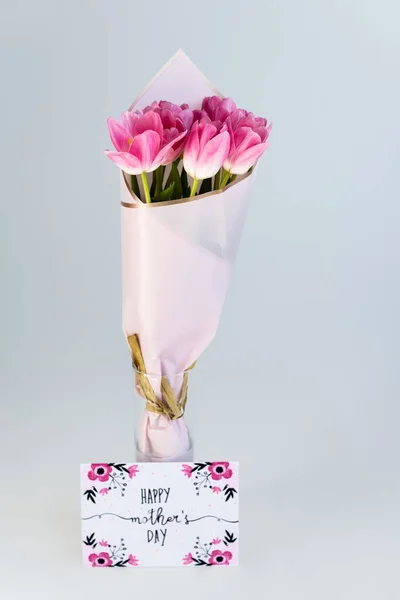 Tulipanes rosados en jarrón cerca de la tarjeta de felicitación con letras felices del día de las madres aisladas en gris - foto de stock