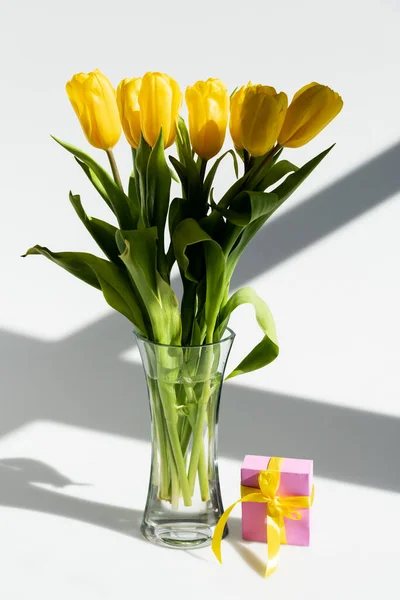 Sonnenlicht auf gelben Tulpen in der Vase nahe Geschenkbox auf weißem, Muttertagskonzept — Stockfoto