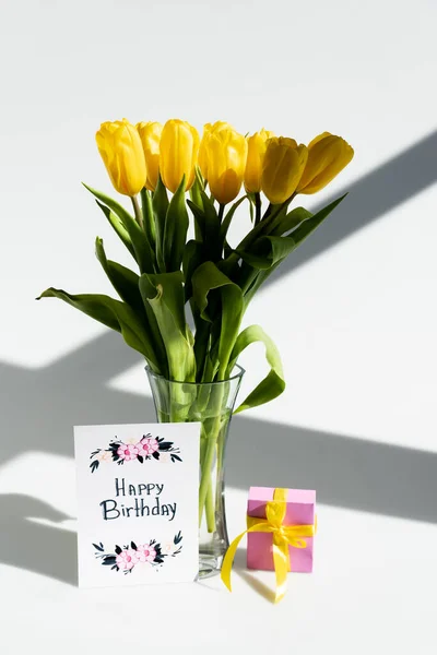 Lumière du soleil sur les tulipes jaunes dans un vase près de la boîte cadeau et carte de vœux avec lettrage joyeux anniversaire sur blanc — Photo de stock