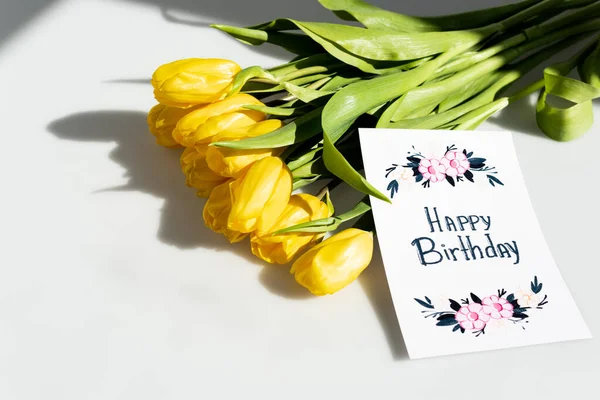 Sunlight on yellow tulips near greeting card with happy birthday lettering on white — Stock Photo