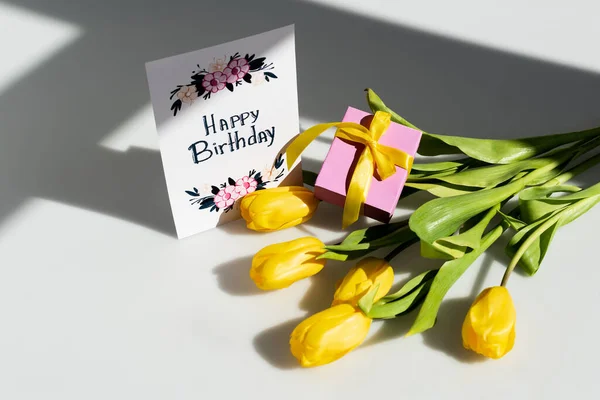 Сонячне світло на жовтих тюльпанах біля подарунку та вітальні листівки з днем народження на білому — стокове фото