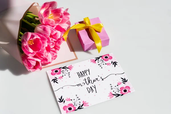 Luz del sol en tulipanes rosados cerca de la caja de regalo y tarjeta de felicitación con letras felices del día de las madres en blanco - foto de stock