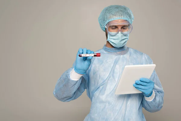 Médico sosteniendo tubo de ensayo con muestra de sangre y letras de coronavirus mientras usa tableta digital aislada en gris - foto de stock