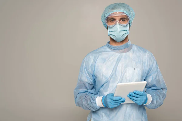 Doctor en googles protectores y máscara médica sosteniendo tableta digital y mirando a la cámara aislada en gris - foto de stock