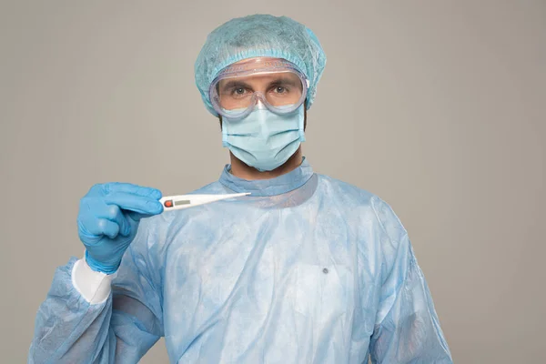 Doctor en googles protectores y máscara médica que sostiene el termómetro aislado en gris - foto de stock