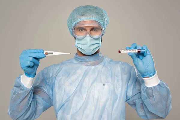 Doctor in medical mask holding thermometer and test tube with blood sample and coronavirus lettering isolated on grey — Stock Photo